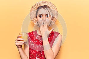 Young beautiful blonde woman wearing summer hat drinking glass of orange juice covering mouth with hand, shocked and afraid for