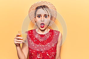 Young beautiful blonde woman wearing summer hat drinking glass of orange juice scared and amazed with open mouth for surprise,