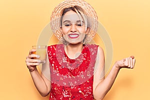 Young beautiful blonde woman wearing summer hat drinking glass of orange juice screaming proud, celebrating victory and success