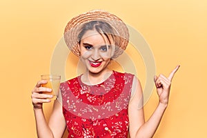 Young beautiful blonde woman wearing summer hat drinking glass of orange juice smiling happy pointing with hand and finger to the