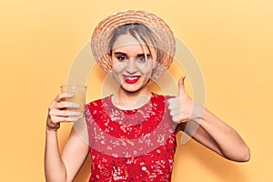 Young beautiful blonde woman wearing summer hat drinking glass of orange juice smiling happy and positive, thumb up doing