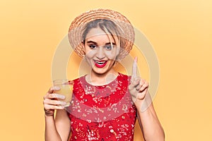 Young beautiful blonde woman wearing summer hat drinking glass of orange juice smiling with an idea or question pointing finger