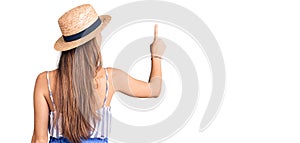 Young beautiful blonde woman wearing summer hat posing backwards pointing ahead with finger hand