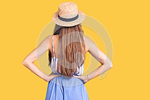 Young beautiful blonde woman wearing summer hat standing backwards looking away with arms on body