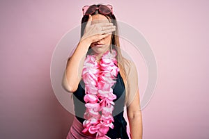 Young beautiful blonde woman wearing swimsuit and floral Hawaiian lei over pink background covering eyes with hand, looking