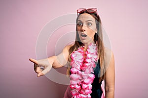 Young beautiful blonde woman wearing swimsuit and floral Hawaiian lei over pink background Pointing with finger surprised ahead,