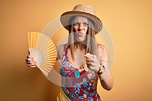 Young beautiful blonde woman wearing swimsuit and summer hat holding fan over yellow background annoyed and frustrated shouting