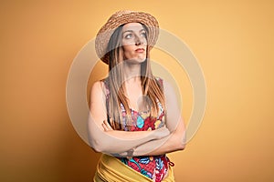 Young beautiful blonde woman wearing swimsuit and summer hat over yellow background looking to the side with arms crossed