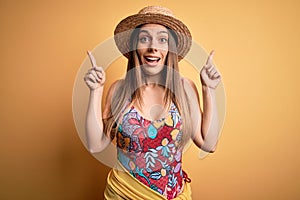 Young beautiful blonde woman wearing swimsuit and summer hat over yellow background smiling amazed and surprised and pointing up