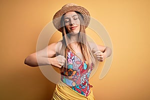 Young beautiful blonde woman wearing swimsuit and summer hat over yellow background stretching back, tired and relaxed, sleepy and