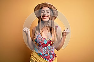 Young beautiful blonde woman wearing swimsuit and summer hat over yellow background very happy and excited doing winner gesture