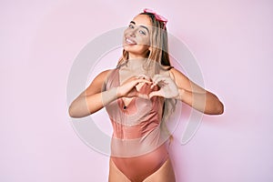 Young beautiful blonde woman wearing swimsuit and sunglasses smiling in love showing heart symbol and shape with hands