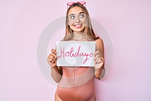 Young beautiful blonde woman wearing swimwear and holding holidays banner smiling looking to the side and staring away thinking