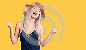 Young beautiful blonde woman wearing swimwear and summer hat very happy and excited doing winner gesture with arms raised, smiling