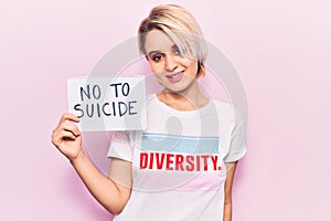 Young beautiful blonde woman wearing t shirt with diversity message holding no to suicide paper looking positive and happy