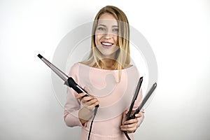 Young beautiful blonde woman wearing trendy pink dress holding curling iron and hair streightener on isolated white