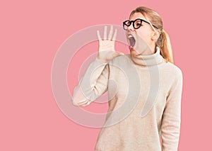 Young beautiful blonde woman wearing turtleneck sweater and glasses shouting and screaming loud to side with hand on mouth