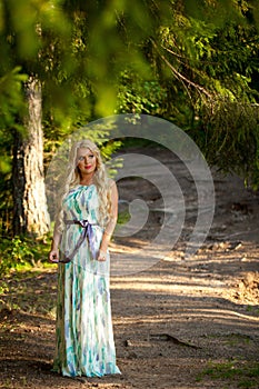 Young beautiful blonde woman in white dress with blue and green flowers posing in forest