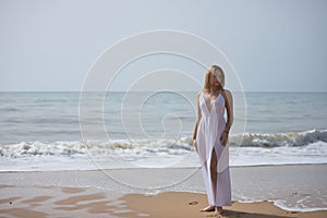 Young, beautiful, blonde woman in white dress, posing on a lonely beach, relaxed and calm. Concept peace, tranquility, solitude,