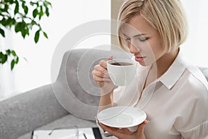 Young beautiful blonde woman in white shirt and glasses is sitting on the sofa in bright office against the window