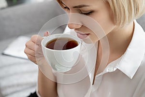 Young beautiful blonde woman in white shirt and glasses is sitting on the sofa in bright office against the window