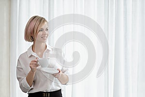 Young beautiful blonde woman in white shirt and glasses is sitting on the sofa in bright office against the window