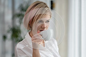 Young beautiful blonde woman in white shirt and glasses is sitting on the sofa in bright office against the window