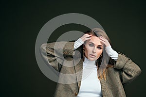 A young beautiful blonde woman in a white turtleneck and a man`s jacket poses on a green olive background in the studio.