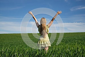 Young, beautiful, blonde woman in a yellow dress, arms raised in the middle of a green wheat field, seen from the back, alone,