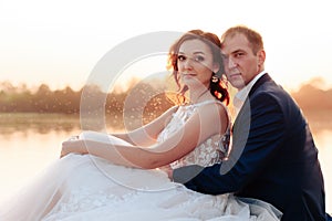 Young beautiful bride and groom resting on the pier near the river at sunset