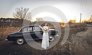 Young beautiful bride is next near to black retro car sunset background. Wedding with vintage old retro car. woman holds beautiful