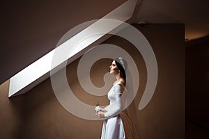 A young beautiful bride is waiting for the groom by the window in a hotel room with a boutonniere in her hands. Wedding morning