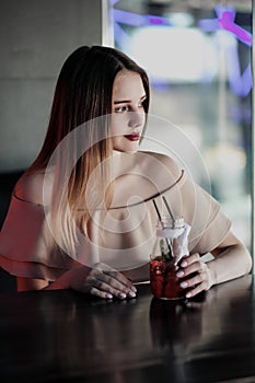 Young beautiful brown-haired girl in a nightclub with neon lights sits by the glass and drinks a red cocktail.