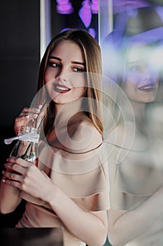 Young beautiful brown-haired girl in a nightclub with neon lights sits by the glass and drinks a red cocktail.