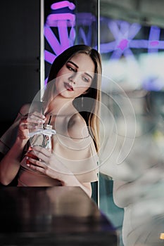 Young beautiful brown-haired girl in a nightclub with neon lights sits by the glass and drinks a red cocktail.