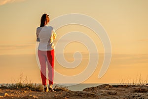 Young beautiful brunete woman in a white jacket and red pants, standing on a cliff over the sea and wait for a call. back view.