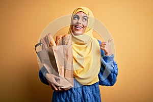 Young beautiful brunette arab woman wearing islamic hijab holding paper bag with bread pointing and showing with thumb up to the