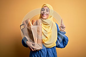 Young beautiful brunette arab woman wearing islamic hijab holding paper bag with bread surprised with an idea or question pointing