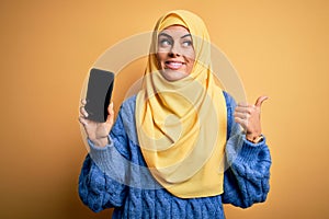 Young beautiful brunette arab woman wearing islamic hijab holding smartphone showing screen pointing and showing with thumb up to