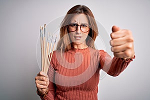 Young beautiful brunette artist woman holding painter brushes over white background annoyed and frustrated shouting with anger,