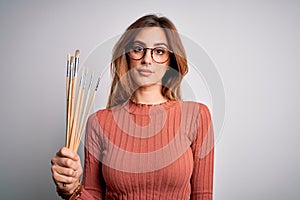 Young beautiful brunette artist woman holding painter brushes over white background with a confident expression on smart face