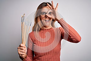 Young beautiful brunette artist woman holding painter brushes over white background with happy face smiling doing ok sign with