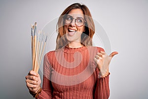 Young beautiful brunette artist woman holding painter brushes over white background pointing and showing with thumb up to the side