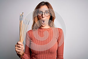 Young beautiful brunette artist woman holding painter brushes over white background scared in shock with a surprise face, afraid