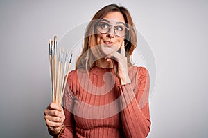 Young beautiful brunette artist woman holding painter brushes over white background serious face thinking about question, very