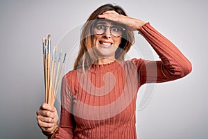 Young beautiful brunette artist woman holding painter brushes over white background stressed with hand on head, shocked with shame