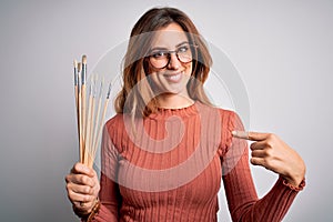 Young beautiful brunette artist woman holding painter brushes over white background with surprise face pointing finger to himself