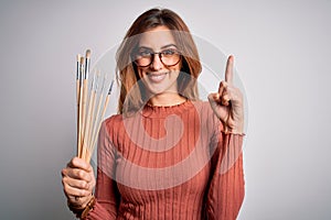 Young beautiful brunette artist woman holding painter brushes over white background surprised with an idea or question pointing