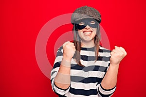Young beautiful brunette burglar woman wearing cap and mask over isolated red background celebrating surprised and amazed for