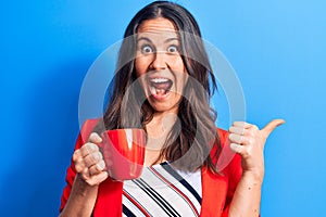 Young beautiful brunette businesswoman drinking mug of coffee over isolated blue background pointing thumb up to the side smiling